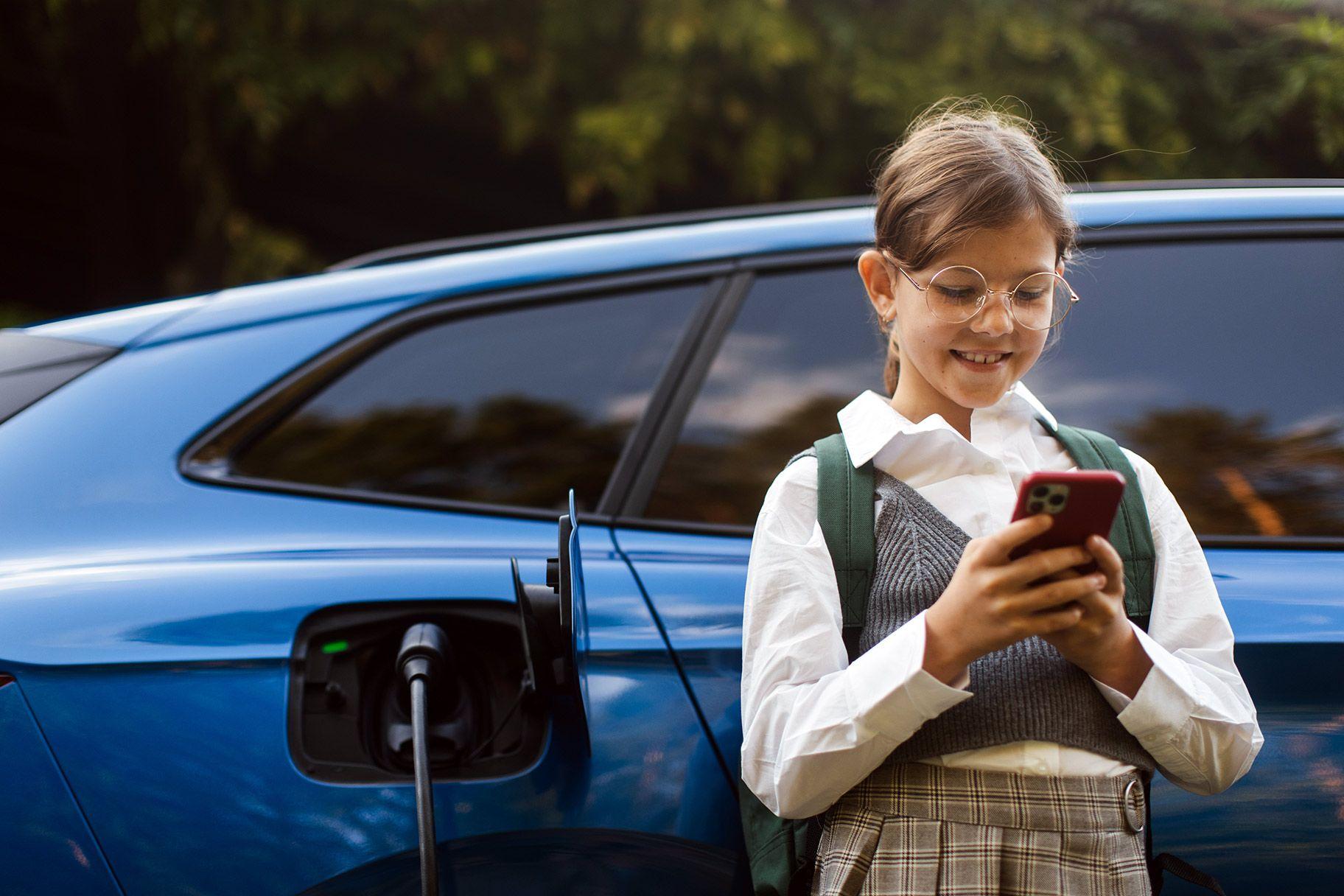 Mädchen mit Smartphone lehnt an ladendem E-Auto